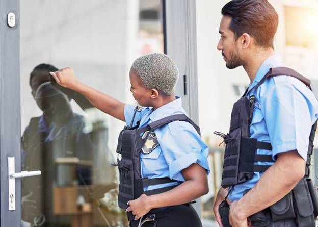 Two police officers stand at a door. The officer on the left knocks on the door, with the other officer to the right.